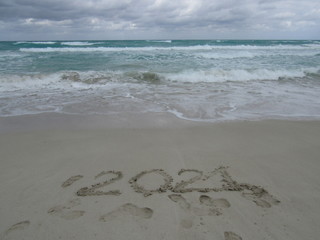 Happy New Year 2021, lettering on the beach with wave and clear blue sea. Numbers 2021 year on the sea shore, message handwritten in the golden sand on beautiful beach background. New Years concept.