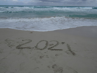 Happy New Year 2021, lettering on the beach with wave and clear blue sea. Numbers 2021 year on the sea shore, message handwritten in the golden sand on beautiful beach background. New Years concept.