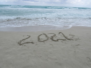 Happy New Year 2021, lettering on the beach with wave and clear blue sea. Numbers 2021 year on the sea shore, message handwritten in the golden sand on beautiful beach background. New Years concept.