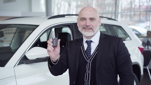 Portrait of happy senior Caucasian man showing car keys at camera and smiling. Confident rich businessman buying new white vehicle in dealership. Business, joy, wealth, success.