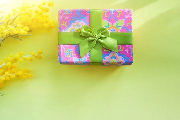 gift bright box with a green bow on a yellow background with a branch of mimosa in the sun. concept of festive spring mood.