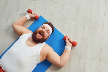 Funny fat man doing exercises with dumbbells on a workout on the floor in the house.