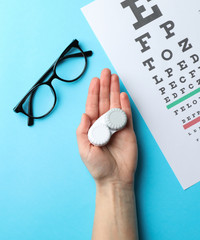 Hand with contact lenses, glasses and eye test chart on blue background, top view