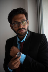 Indian Bengali tall, dark, handsome brunette young man in office wear is standing in front of a glass window adjusting his spectacle in a corporate office/bpo/call center. Indian corporate lifestyle.