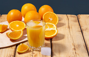 A glass of freshly squeezed orange juice with ice cubes and a wooden board with oranges on a wooden table. Healthy eating concept