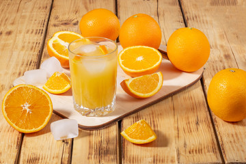 A glass of orange juice with ice cubes and a wooden board with oranges on a wooden table. Fresh orange juice.