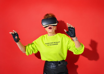 Forever Young! Elderly woman in bright outfit with glasses of virtual reality, touching something on red background