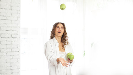Happy woman with curly hair playing with green apple over white background at home