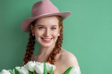 Spring beauty, fashion concept: close up studio portrait of young happy smiling natural redhead girl with freckled skin, wearing pink hat, earrings, holding white tulips. Green mint color background