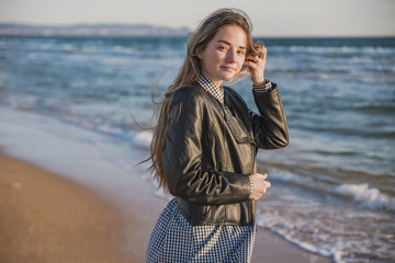 a pretty girl in a plaid dress and jacket walks along the sandy beach