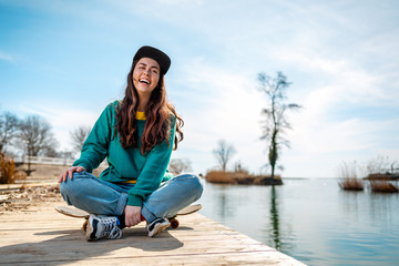 A young Caucasian hipster woman sits cross-legged on a skateboard and laughs merrily. In the background, the sea and the coastline. Concept of sports lifestyle and street cultures