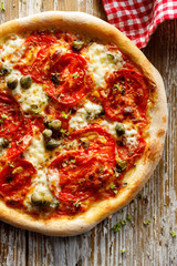 Homemade tomato pizza with addition mozzarella cheese  and capers on a rustic wooden board, top view