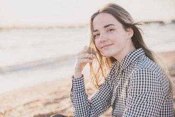 a pretty girl in a plaid dress and jacket sits on the sandy beach at sunset