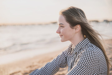 a pretty girl in a plaid dress and jacket sits on the sandy beach at sunset