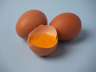 Two whole raw brown chicken eggs and a broken half egg with yellow yolk closeup on a blue background.