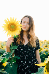 a young beautiful girl in a black floral dress stands in a field among sunflower flowers at sunset