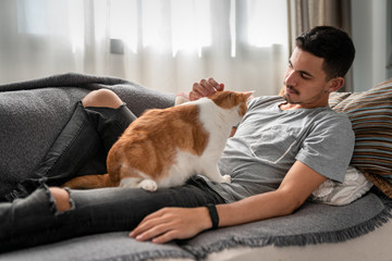 hombre joven recostado en un sofa, acaricia a un gato blanco y marron sentado encima de el