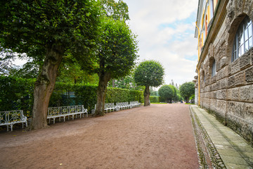 An empty dirt path around Catherine's Palace lined with trees, landscaping and white benches in...