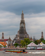 Naklejka premium A colorful long tail boat sails on the Chao Phraya river in front of the Wat Arun temple, against cloudy sky, Bangkok, Thailand