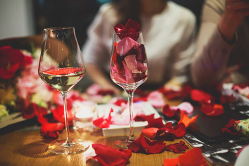 Beautiful glass with rose leafs, rose-petal, rose petals on a romantic date with wine, candles in candlelight and with celebrating couple on the background, interior for a romantic date diner