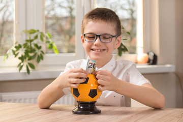 Chłopiec w wieku szkolnym siedzi przy stole i szeroko się uśmiecha. Wkłada do skarbonki w kształcie piłki nożnej banknot o nominale 20 euro.
