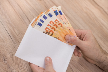Męska dłoń trzyma białą kopertę i wkłada do niej banknoty Euro o nominałach 50 Euro.