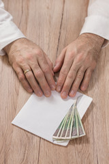 Mężczyzna w białej koszuli trzyma końcami palców białą kopertę wypełnioną banknotami  polskiej walucie PLN  i przesuwa ją po stole dając łapówkę. 