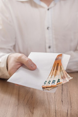 Męska dłoń trzyma białą kopertę wypełnioną banknotami waluty Euro i podaje ją ponad stołem.