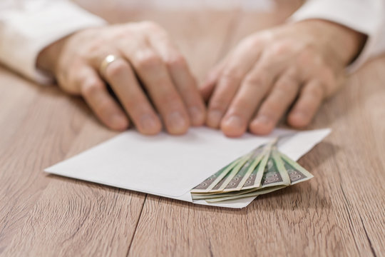 Pieniądze, banknoty w polskiej walucie polski złoty leżą na stole w kopercie. Na drugim planie męskie dłonie trzymają palcami r&oacute;g koperty.