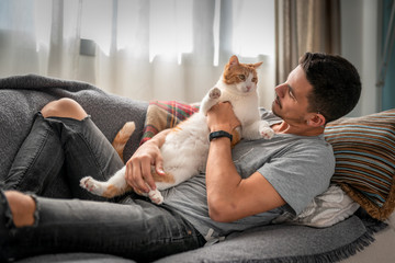 hombre joven recostado en un sofa, toma entre sus brazos a un gato blanco y marron y lo mira con...