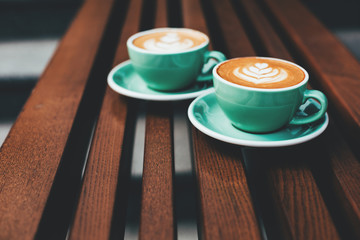 Two cups of cappuccino with latte art on wooden background. Beautiful foam, greenery ceramic cups, stylish toning, place for text.