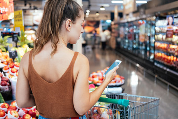 Back view of young woman holding a phone with a shopping list stock photo. Woman using mobile phone while shopping in supermarket, vegetable department store