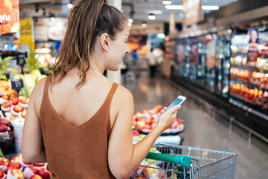  Smiling woman shopping healthy food in supermarket stock photo. Female hands buy nature products using smart phone in vegetable department store