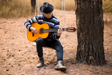 young handsome attractive man in sweater playing guitar in forest sitting on swing. Casual man, life style.