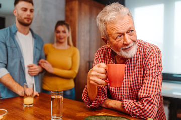 Elderly man with grey hair and beard is drinking coffee. His son and daughter are standing next to him.