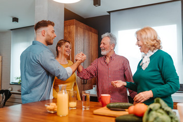 Young man and his father are shaking hands while standing in the kitchen. Vegetables and squeezed juice are on the kitchen table.