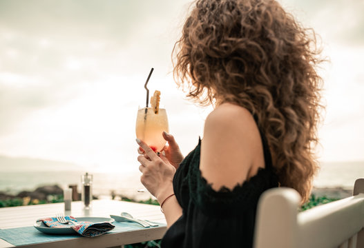 Closeup portrait. Stylish woman with curly hairstyle having fun at luxury summer bar. Overwhelming girl in black sexy dress with colorfull cocktail. Lady relaxing on sunset .