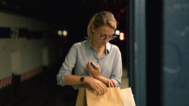 Millennial female passenger in trendy optical glasses searching purchases in paper shopping bags while waiting for subway metro on public station, charming woman in spectacles checking buy stuff