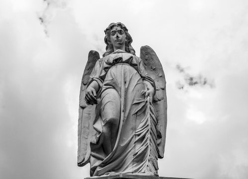 An old angel sculpture looking down in low angle shooting in top of mausoleum cemetery. Blue and cloudy sky at background. Black and White photography