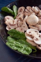 whole and sliced ​​mushrooms, on black plate with green spinach leaves, on blue background