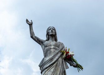 Fototapeta premium An old Jesus Christ resurrected statue with a multicolor bouquet flowers in a hand and a blue cloudy sky at background
