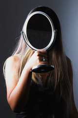 Unrecognizable, young woman holding a mirror in her hand and covering her face. Girl on a grey and...