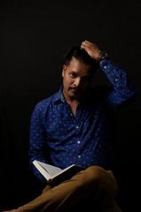 Portrait of a handsome and intelligent Indian brunette man wearing blue shirt with white stars sitting on a chair with a book in his hands. Indian lifestyle and fashion portrait