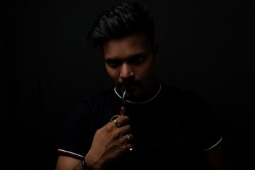 Close up portrait of a handsome and intelligent Indian brunette man wearing a solid black t shirt standing before a copy space black background holding a tobacco. Indian lifestyle and fashion portrait