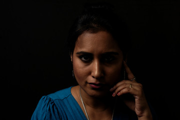 Fototapeta na wymiar Close up face portrait of a beautiful Indian Bengali brunette woman in light and shadow before a black copy space background wearing a blue top. Indian lifestyle and fashion portrait