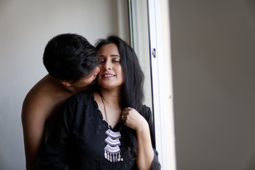 Attractive Indian Bengali brunette couple sharing intimate moments in bedroom in front of white background while the man trying to kiss the girl standing in front of a window.  Indian lifestyle 