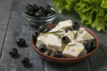 Lettuce leaves and a bowl of feta cheese and olives on a black wooden table.