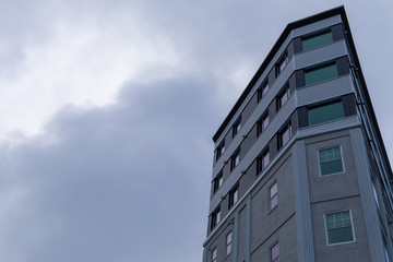 An upward partial view of a tall concrete building with lots of double hung windows.  A grey cloudy sky is the background to the condo building. The textured exterior walls are of two  tones of grey. 