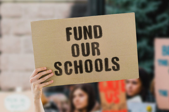 The phrase " Fund our schools " on a banner in men's hand. Human holds a cardboard with an inscription. Social workers. Government. Education. Governmental budget. Money. Sponsoring. Future