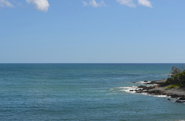 Fototapeta premium A calm blue ocean and a blue sky meet at the horizon. A rocky headland with some trees is to one side. There are some soft white clouds.